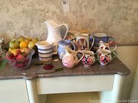 A setting showing 10 pitchers arranged on a table, along with a basket of fruit and a bowl of strawberries and cherries.
