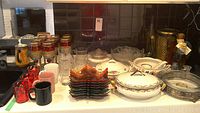 Wide view of lot showing mixed glassware including wine glasses, mugs, stack of leaf-shaped glass plates, and various porcelain serving dishes with lids and floral patterns.