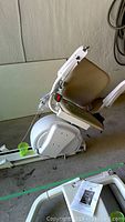 Photo showing side view of beige cushioned chair lift mounted on a white mechanical rail system with power cord and control buttons on armrest.