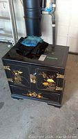 Front view of the Asian inspired black lacquered side table with floral bird artwork on cupboard doors and drawer front, showing the hinged doors and brass hardware.