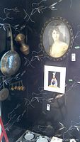 Copper pots and hanging utensils displayed on black wall with decorative silver leaf pattern, an oval framed portrait print above.