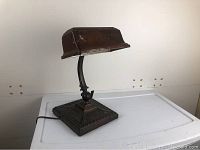 Full view of antique metal desk lamp on white surface showing curved arm and ornate square base.
