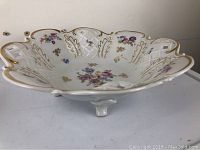 Front and top angled view of the white porcelain open work fruit bowl showing floral decoration, gold edging, and scalloped rim.