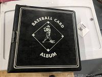 Photo of closed black baseball card album with 'Baseball Card Album' and batter graphic on the cover.
