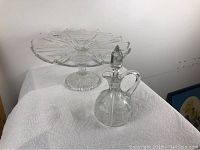 Photo of the clear glass pedestal cake plate and cruet together on a white cloth background showing full shapes and outlines.