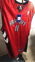 Full front view of a red Toronto Raptors basketball jersey with black and white side panels, hanging showing original tags.