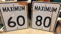 Two metal maximum speed limit signs for 60 and 80 speed limits, showing front face with black text on white background with rust and wear.
