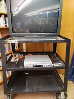 Front view of the JVC TV on a black metal cart with shelves, showing the TV screen and placement of the combo player.