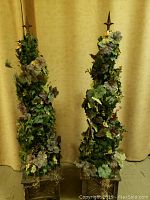 Pair of tall artificial topiary floral arrangements with green and muted foliage and square bases in front of yellow drape background.