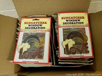 Photo of two stacks of 24 boxed suncatcher window decorations in a cardboard box, showing the 'LOVE' rainbow design and packaging