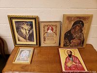 Five framed religious pictures arranged on a wooden surface against a beige wall, showing a variety of religious figures and styles.