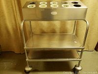 Front view of the stainless steel cafeteria cart showing eight cylindrical serving ware holders on the top and two stainless steel shelves below, mounted on four caster wheels.