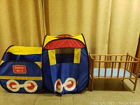 Photo showing the wooden doll crib next to the train-themed foldable fabric play tent with yellow, red, and blue colors.