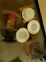 Overview of three white round ceiling speakers including one mounted in orange housing, two spools of wiring (red and black) placed on floor.