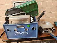 Photo showing blue plastic crate filled with metal trays, pans, pots, and a frying pan with a black handle on a wooden surface.