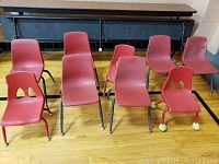 Nine red plastic chairs in a group, seven upright and two shorter wheeled, on a wooden floor with black and wood furniture in background.