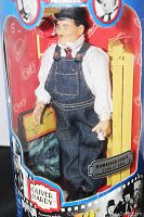 Full view of the Oliver Hardy figurine inside original packaging showing denim overalls, white shirt, red tie, and original box graphics with portraits.