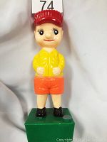 Front view of vintage collectible boy toy doll on green square base wearing red cap, yellow jacket, orange shorts, black shoes