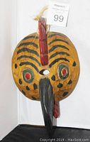 Front view of the wooden bird mask showing painted eyes, stripes, and attached hanging string.