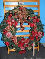 Front view of large Christmas wreath decorated with red flowers, striped bows, and gold accents hung on wooden chair.