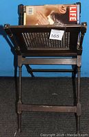 Front view of antique wooden magazine rack showing turned legs and woven panel storage area with a magazine inside.