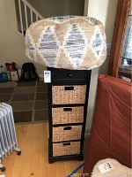 Tall black storage unit with 1 drawer and 4 wicker baskets including pull handles, standing on wooden floor, next to a couch and heater.
