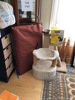 Portable dog crate and pet accessories including scratching posts, feeding dishes, and puppy pad box photographed indoors on hardwood floor beside furniture and stairs.