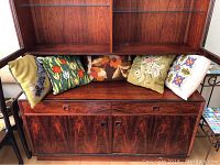 Five needlepoint pillows arranged on a dark wood cabinet, showing all designs and velveteen backing visible on multiple pillows.