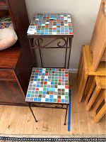 Two wrought iron tile top tables stacked, showing colorful square tiles on top and the rusted wrought iron legs and frame.