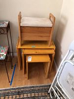 Photo showing three wooden nesting tables stacked and a small wooden storage bench with beige upholstered cushioned top stacked on top of the largest table.