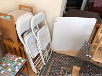 View of white card table leaning against wall with four white metal folding chairs stacked next to it, showing the overall items in the lot.