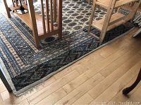 Partial view of the rug under a dining table showing geometric and floral pattern in blue, cream, brown and rust colors. Edges visible with some frayed fringe.