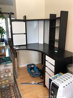 Photo showing black corner desk placed in a corner with built-in shelving and two white storage bins, along with part of the mobile storage unit visible.