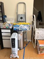 Photo displaying the COLOMBO 3-step ladder behind a DeLonghi heater, the Hurricane Spin Scrubber box on the ladder, and dented stainless steel step-on can beside them.