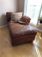 Front-side angle view of brown leather chaise lounge with pillow and blanket near window.