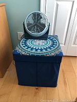 Collapsible fabric storage box with cushioned seat in blue and turquoise mandala pattern with Lasko Breeze Machine fan on top