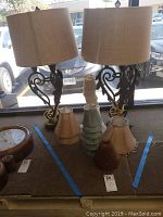 Two ornate metal table lamps with beige drum shades shown on a surface by the window with six additional lamp shades in front