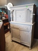 Front view of painted two-piece wood cabinet with ornamental carved panels on upper and lower doors.