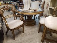 View of two wooden arm chairs with carved backs and beige upholstered seats arranged around a round wooden table with decorative painted details on top.