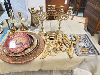 Photo showing multiple brass candlesticks, decorative trays (metal and painted), brass bed warmer, brass pitcher, and gold-painted wood sconces on a laid cloth.