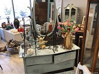 Overview of table with religious statue, silk tulips, angel figurines, and metal racks with other furniture in background