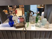 Full lot display showing hat pins in blue vase, pottery pitchers and vase, metal purse, bracelets, perfume and glass bottles on table