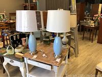 Four table lamps on a wooden table: pair of blue ceramic lamps with different white shades, missing finial on one; beige-shade lamp with carved white base; fourth lamp with damaged painted base.