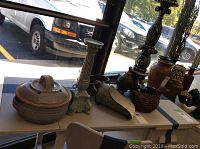Overview of eight decorative items on table including pottery, candlesticks, metal duck, and other metal and ceramic ware in natural light near window