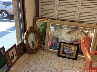 View of multiple framed art pieces on the floor against a wall, showing different sizes and styles, including a large framed painting and an ornate mirror.
