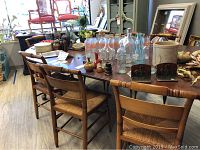 Full view of table top showing dark brown wooden surface, various glass bottles and vases scattered on top, chairs around.