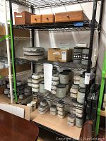 Shelving unit showing multiple jars, tubs, and containers of Artisan Enhancements furniture refinishing paints and products with wooden organizer drawers on top shelf.