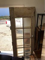First photo showing full view of one weathered wooden door with glass panes, doorknob visible, leaning against a wall near a doorway.