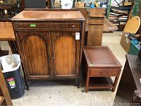 Vintage wooden chest with a single drawer and two paneled cabinet doors. Missing one drawer pull on the top drawer. Positioned next to a wooden side table.