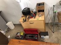 Wide shot showing red toolbox with hand tools, two hammers, mallet, furniture moving pads, unopened paint can, and CD player on wooden cabinet.